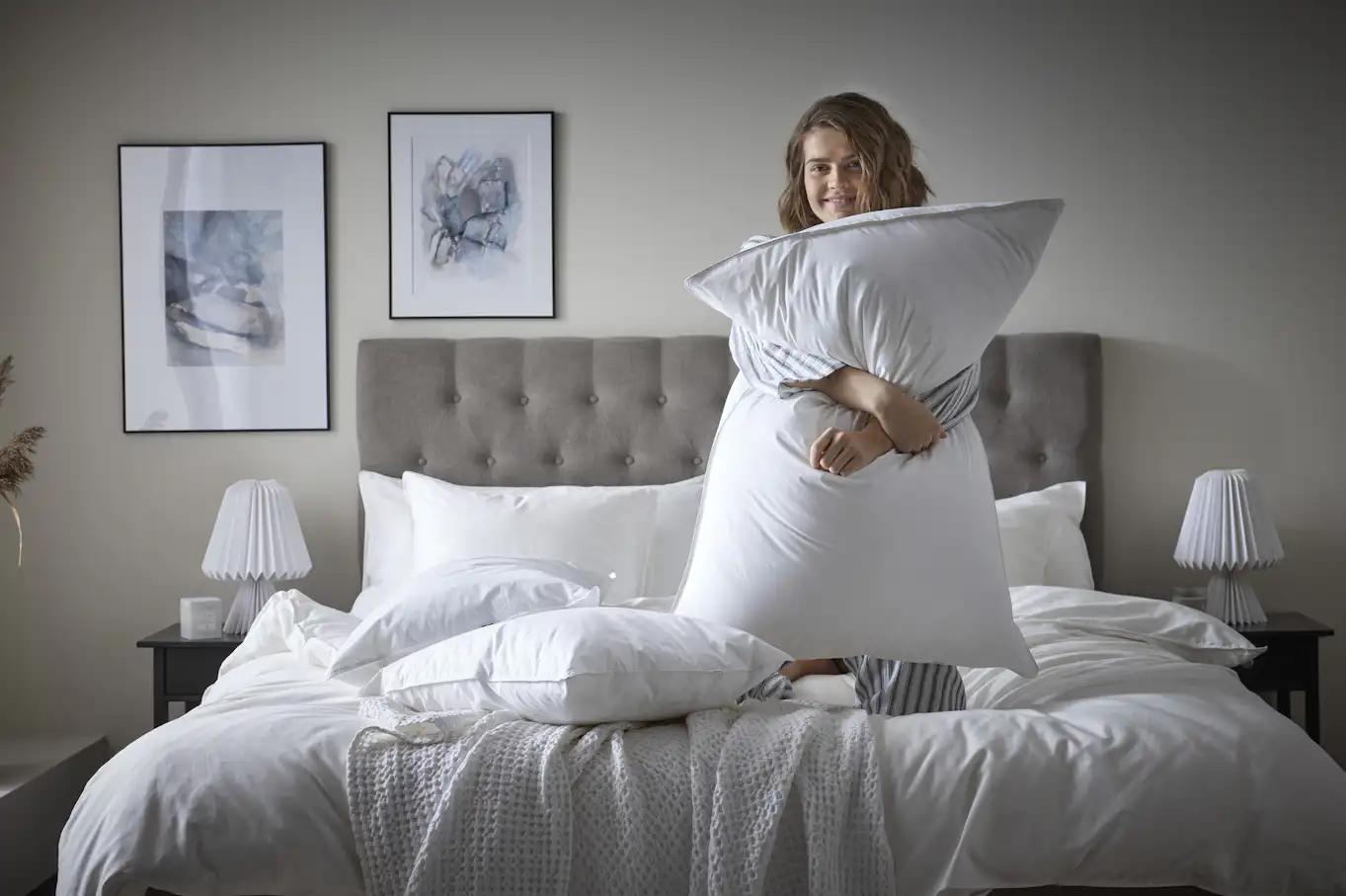 Kvinna i randig pyjamas står på knä i en säng och ler mot kameran och kramar en kudde.