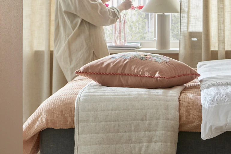 en tjej står vid ett fönster i ett sovrum där det finns en säng med rosa påslakan och ett beiget överkast