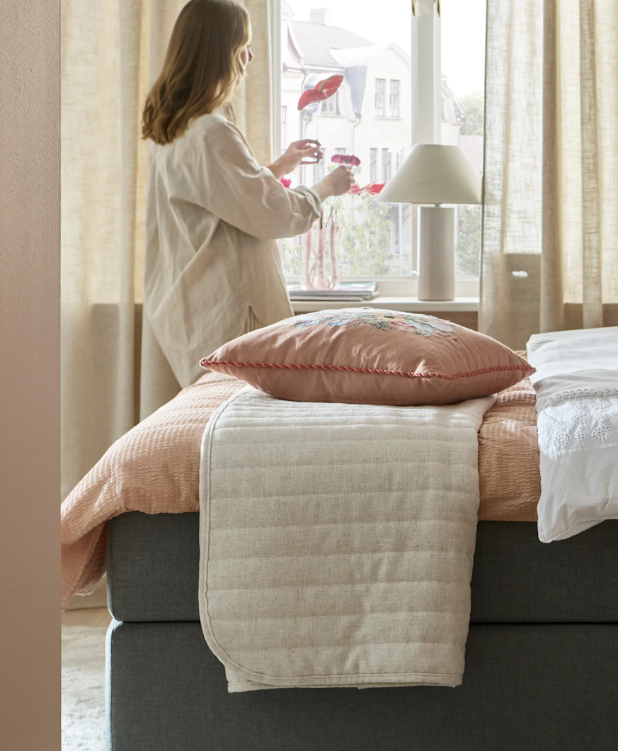 en tjej står vid ett fönster i ett sovrum där det finns en säng med rosa påslakan och ett beiget överkast