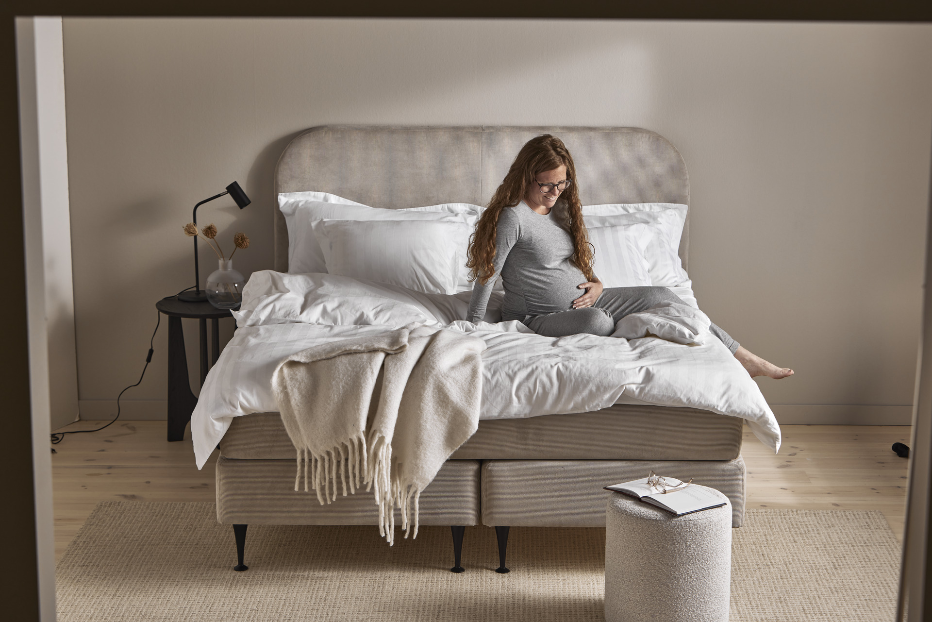 Gravid kvinna i grå pyjamas halvsitter i kontinentalsäng, tittar ner mot magen och håller ena handen på magen.