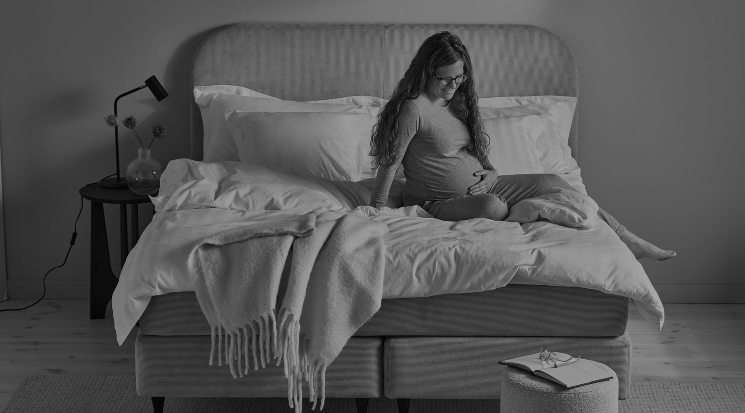Gravid kvinna i grå pyjamas halvsitter i kontinentalsäng, tittar ner mot magen och håller ena handen på magen. Bild i gråskala. 