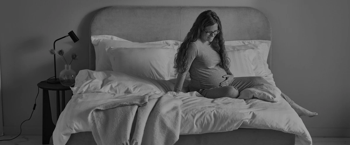 Gravid kvinna i grå pyjamas halvsitter i kontinentalsäng, tittar ner mot magen och håller ena handen på magen. Bild i gråskala.