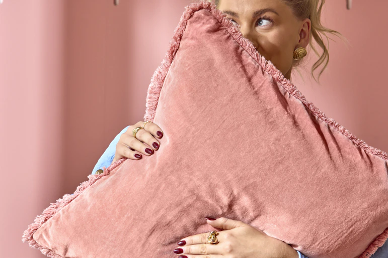 en blond tjej står och håller upp en rosa kudde framför ansiktet