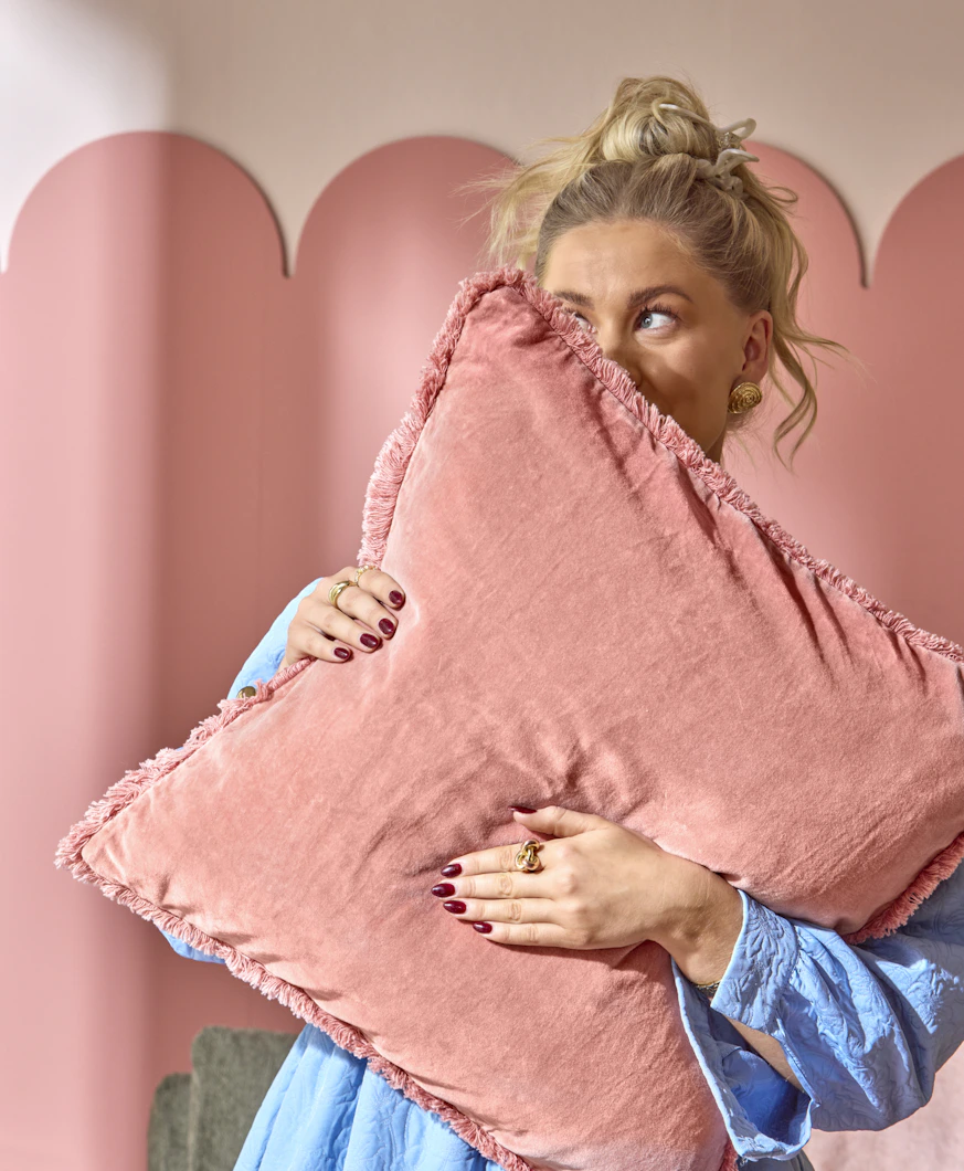 en blond tjej står och håller upp en rosa kudde framför ansiktet