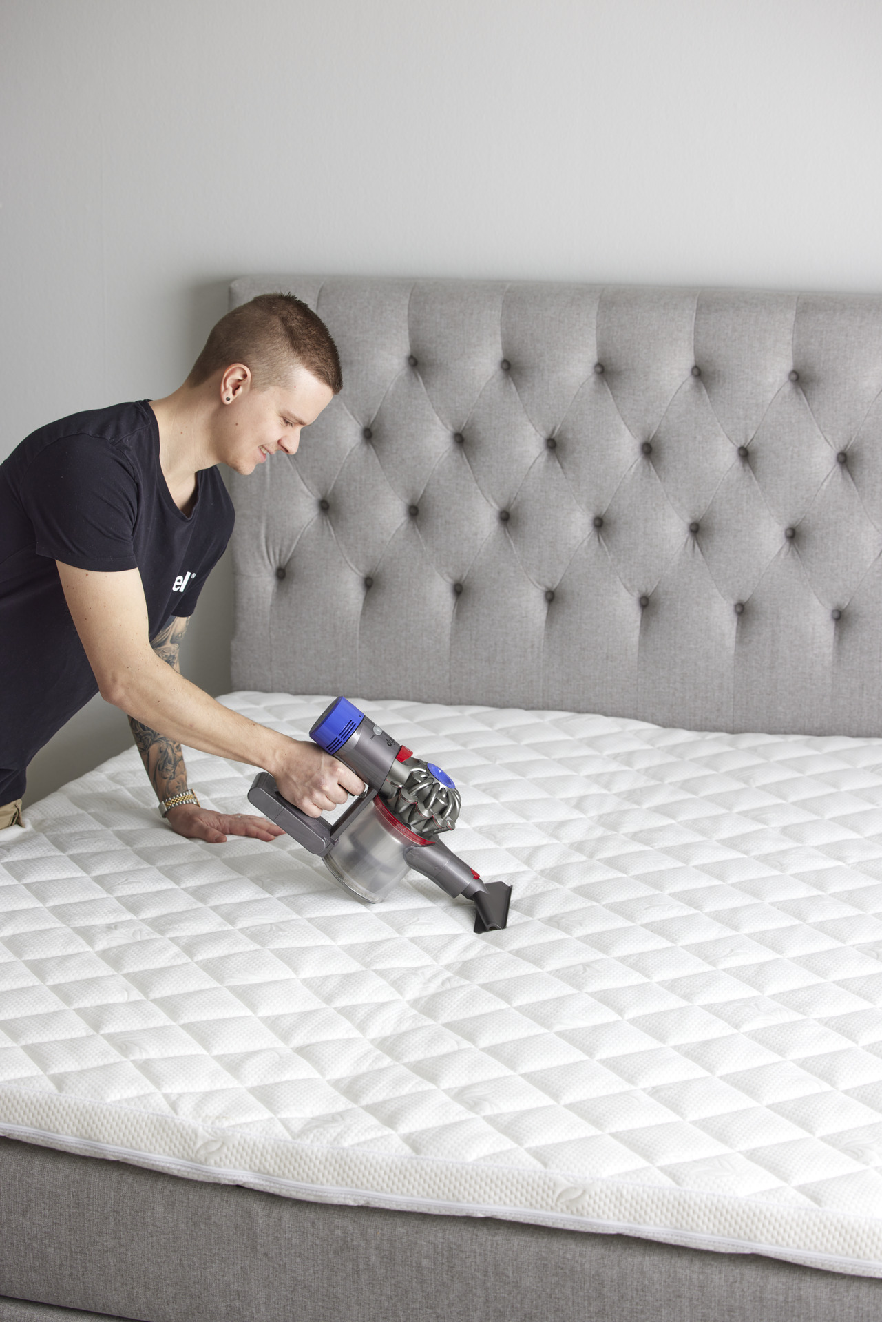 Ung man står lutar sig över en säng utan sängkläder och dammsuger bäddmadrassen 