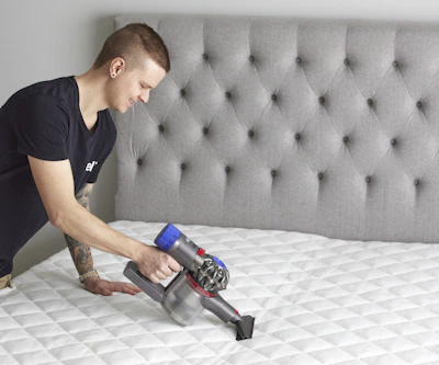 Ung man står lutar sig över en säng utan sängkläder och dammsuger bäddmadrassen