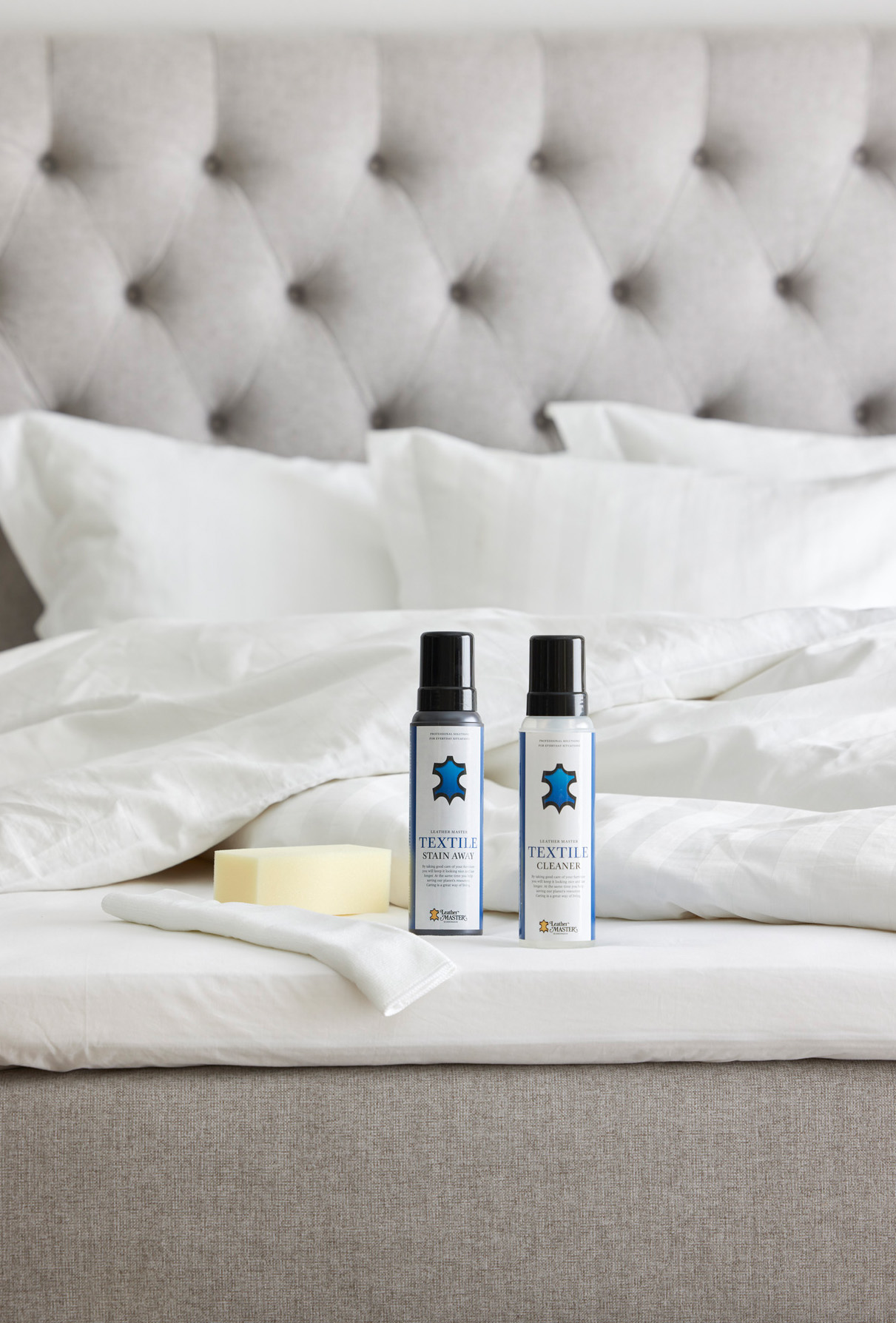 Två flaskor möbelvård med blå logotyper står uppställda på en säng intill en tvättsvamp och fibertrasa. 