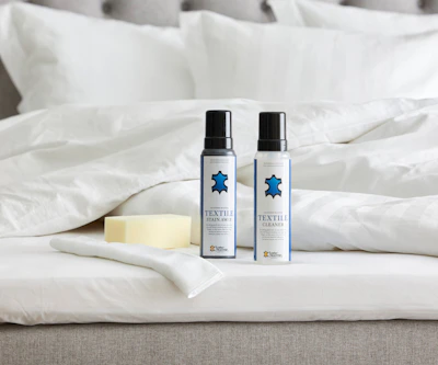 Två flaskor möbelvård med blå logotyper står uppställda på en säng intill en tvättsvamp och fibertrasa.