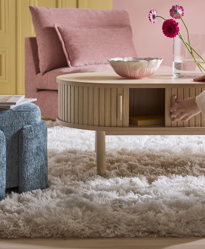 en vit ryamatta ligger på ett golv under ett träfärgat soffbord, en rosa soffa och blå puff