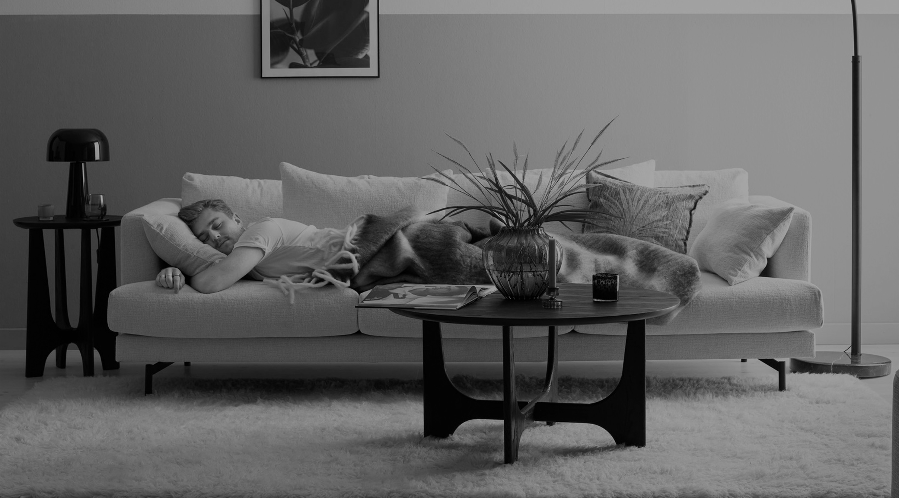 Ung man ligger och sover på mage kramandes en kudde i en soffa i ett vardagsrum. Bild i gråskala. 