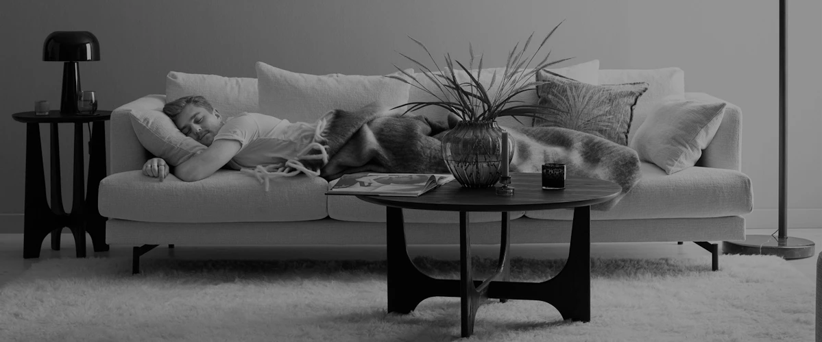 Ung man ligger och sover på mage kramandes en kudde i en soffa i ett vardagsrum. Bild i gråskala.