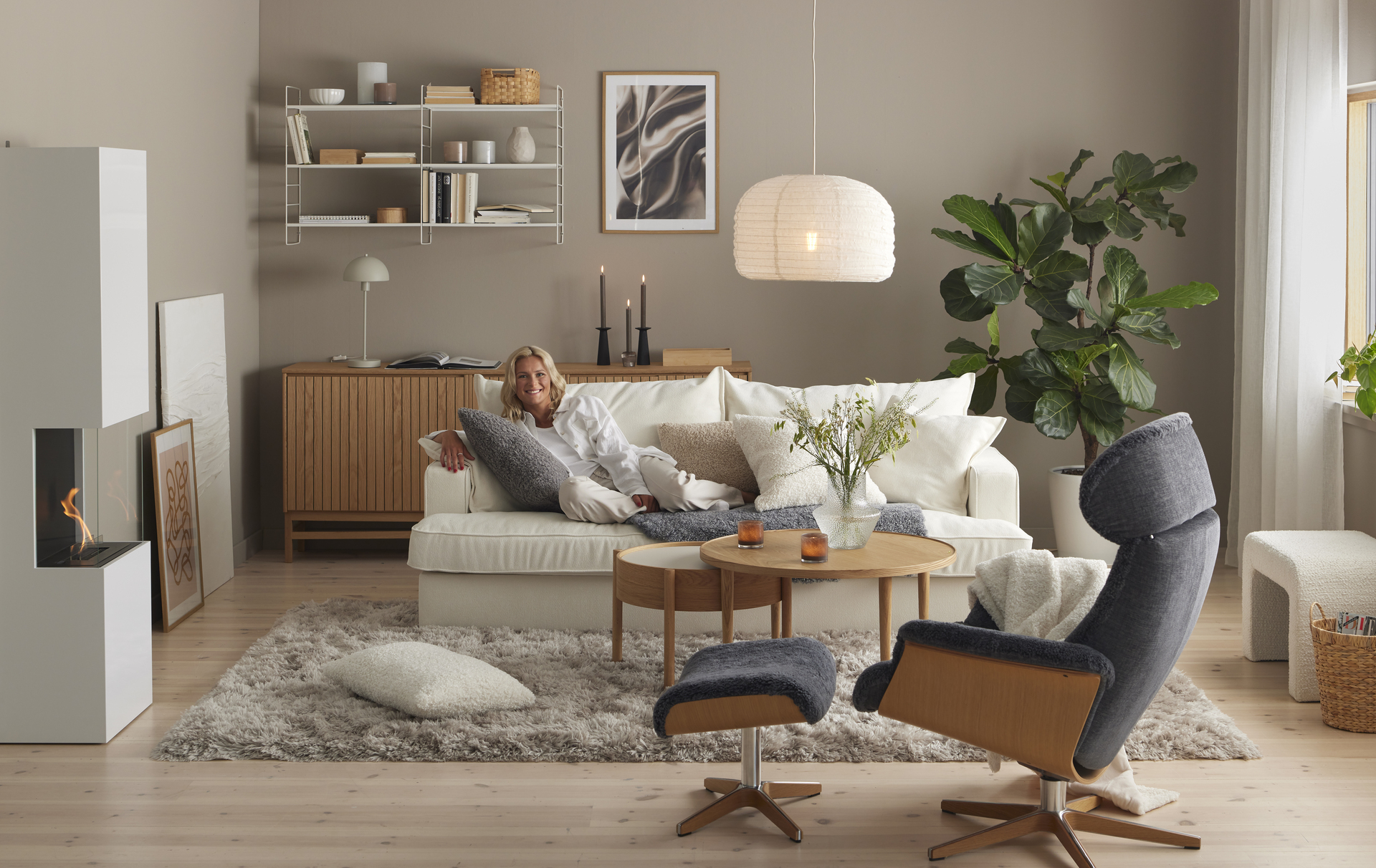 Vardagsrumsmiljö med soffa centrerat placerad i rummet, där skidstjärnan Frida Karlsson sitter i en vit soffa i myskläder och håller om en krullig grå kudde. 