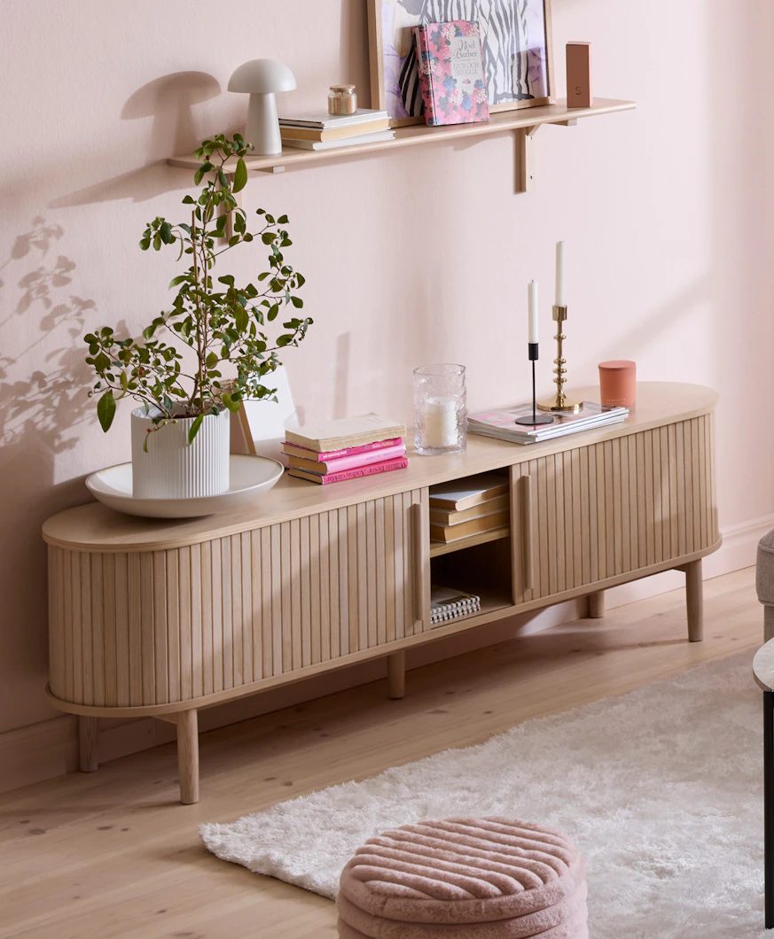 en tv-bänk i ljust trä med ribbor står i ett rum med rosa väggar