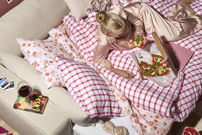 I en bäddsoffa ligger en kvinna i pyjamas på ett rosa rutigt täcke och äter pizza
