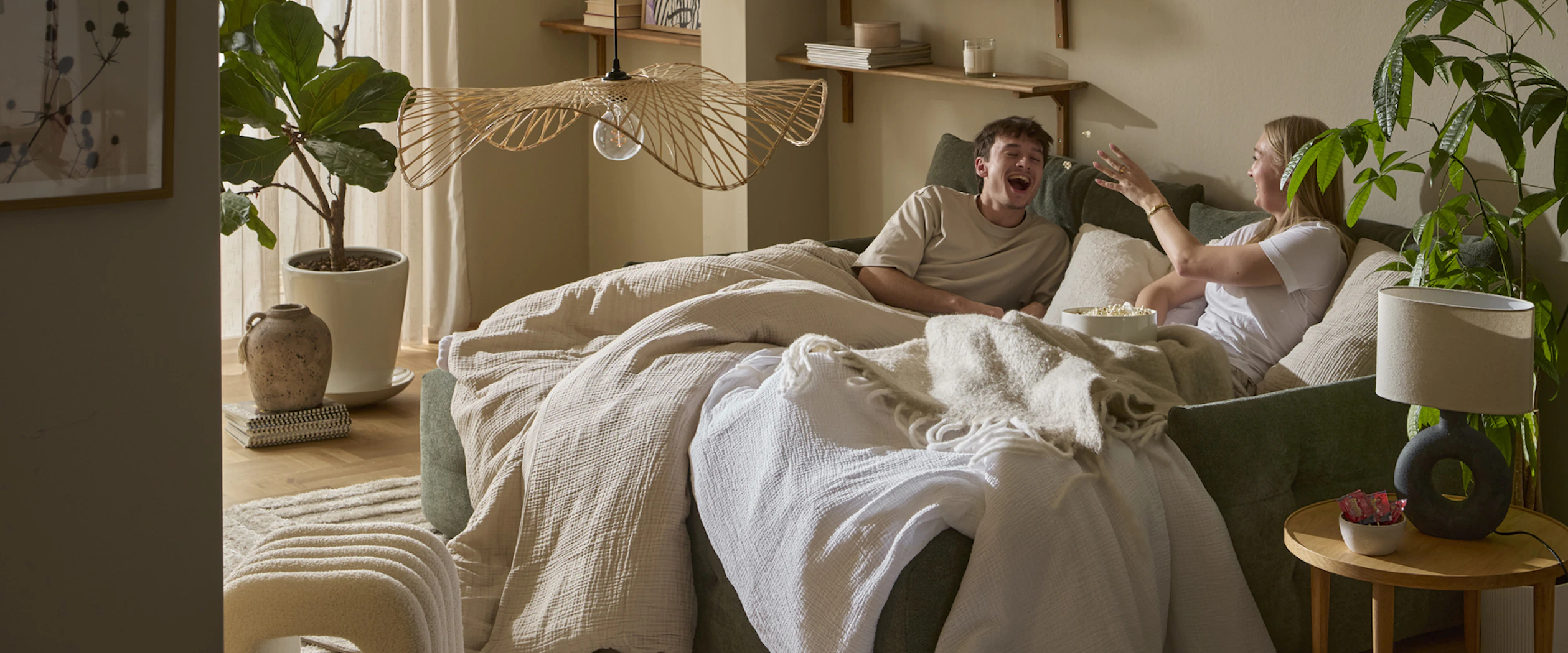 I en grön bäddsoffa sitter en tjej och en kille nedbäddade under ett täcke och kastar popcorn till varandra