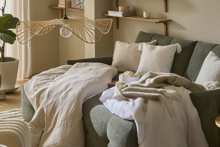 I ett vardagsrum står en grön, modern bäddsoffa. I soffan ligger täcken och kuddar med beiga påslakan och örngott