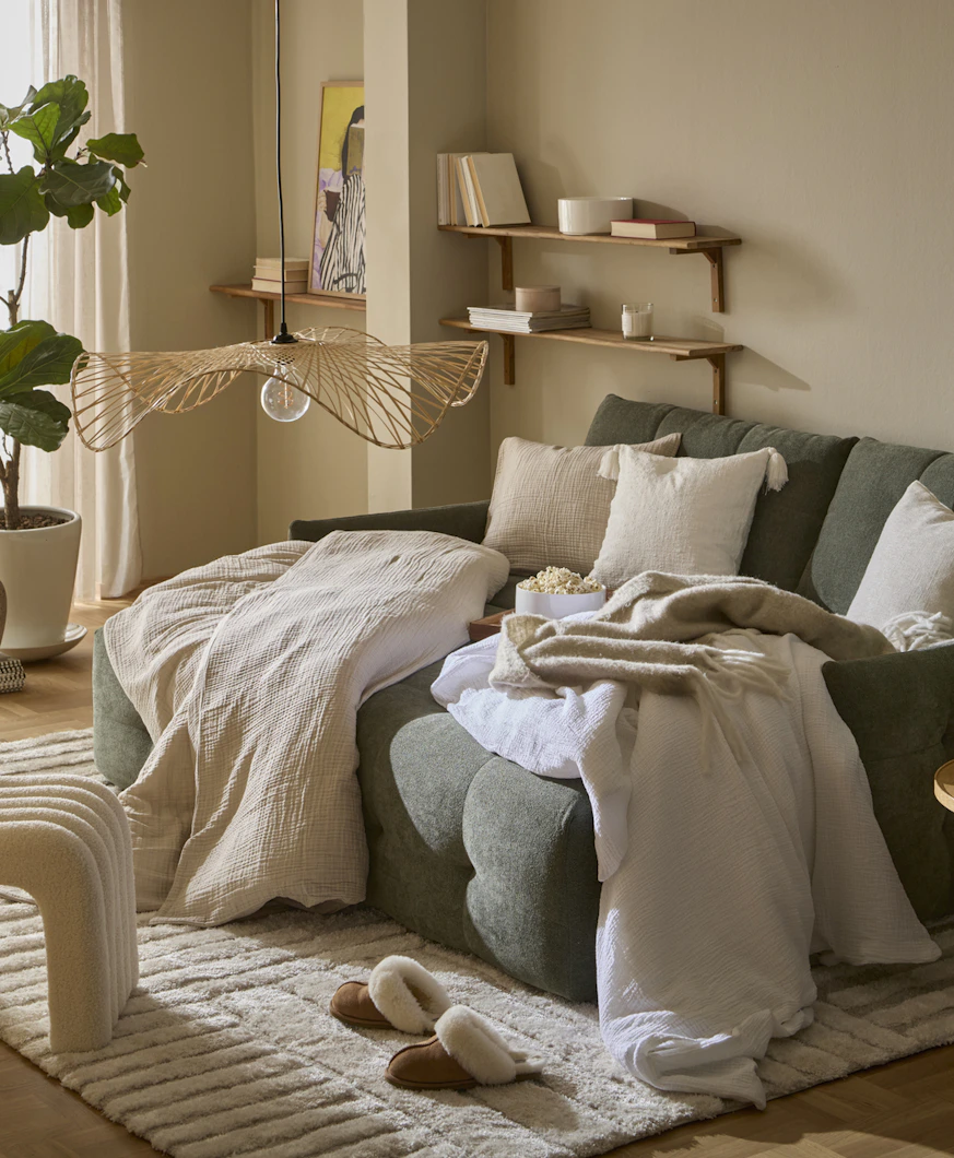 I ett vardagsrum står en grön, modern bäddsoffa. I soffan ligger täcken och kuddar med beiga påslakan och örngott