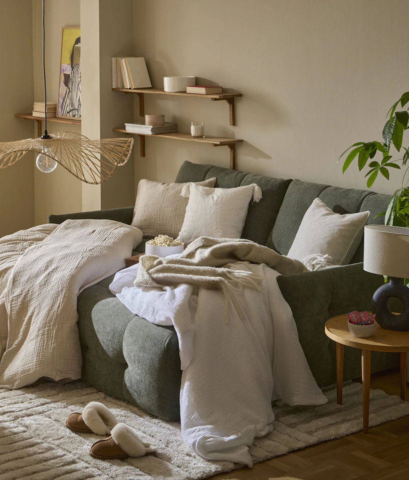 I ett vardagsrum står en grön, modern bäddsoffa. I soffan ligger täcken och kuddar med beiga påslakan och örngott