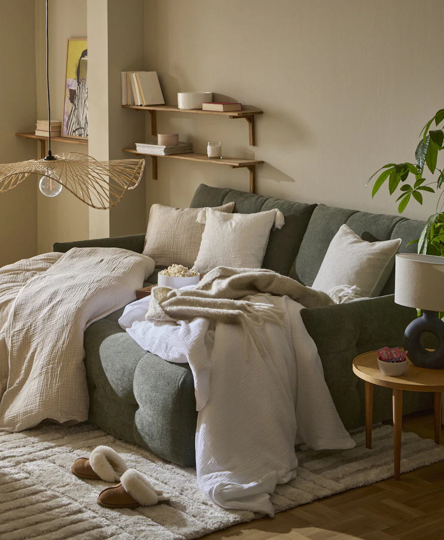 I ett vardagsrum står en grön, modern bäddsoffa. I soffan ligger täcken och kuddar med beiga påslakan och örngott