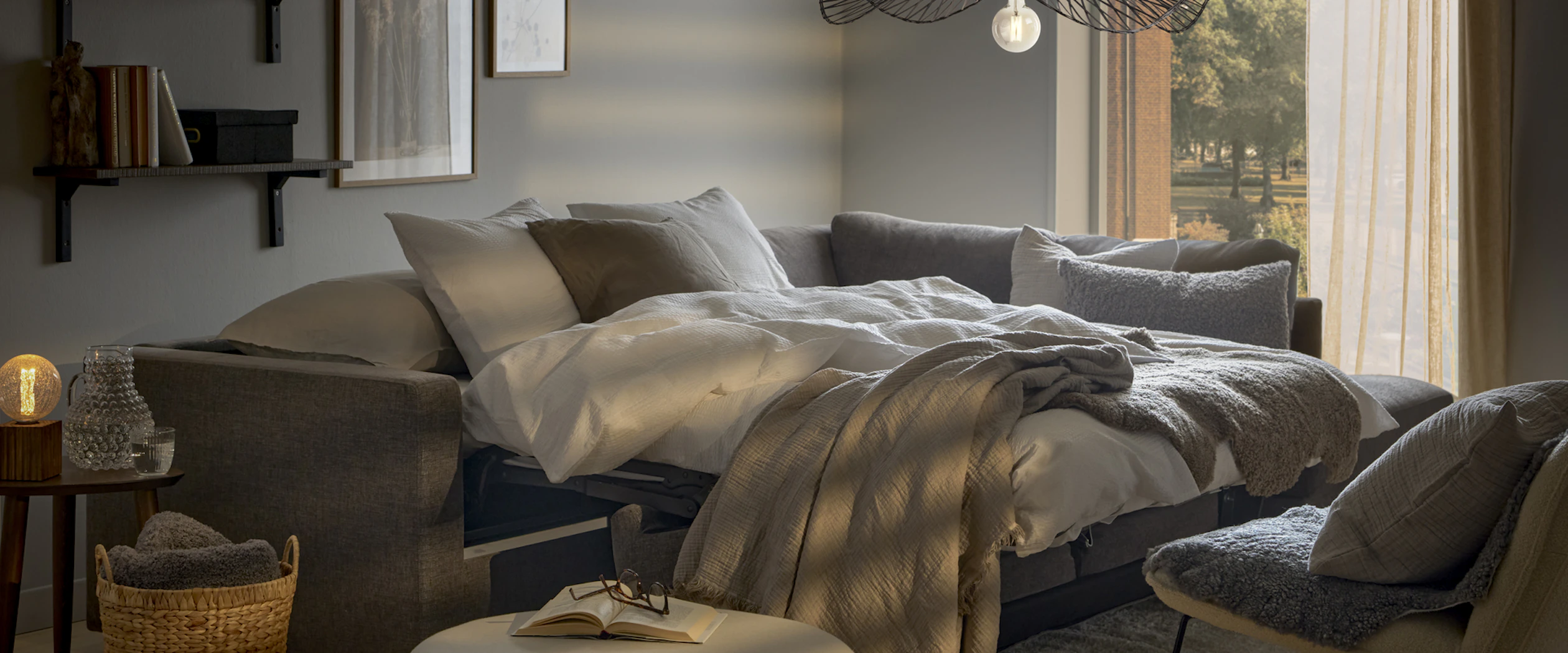 I ett vitt rum står en grå bäddsoffa utbäddad med fluffiga täcken och kuddar