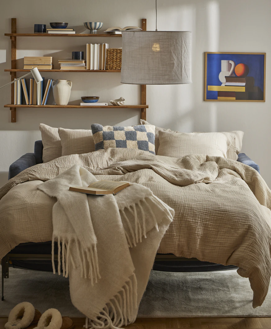 I ett ljust vardagsrum står en utfälld bäddsoffa med sängkläder i beige och blått.