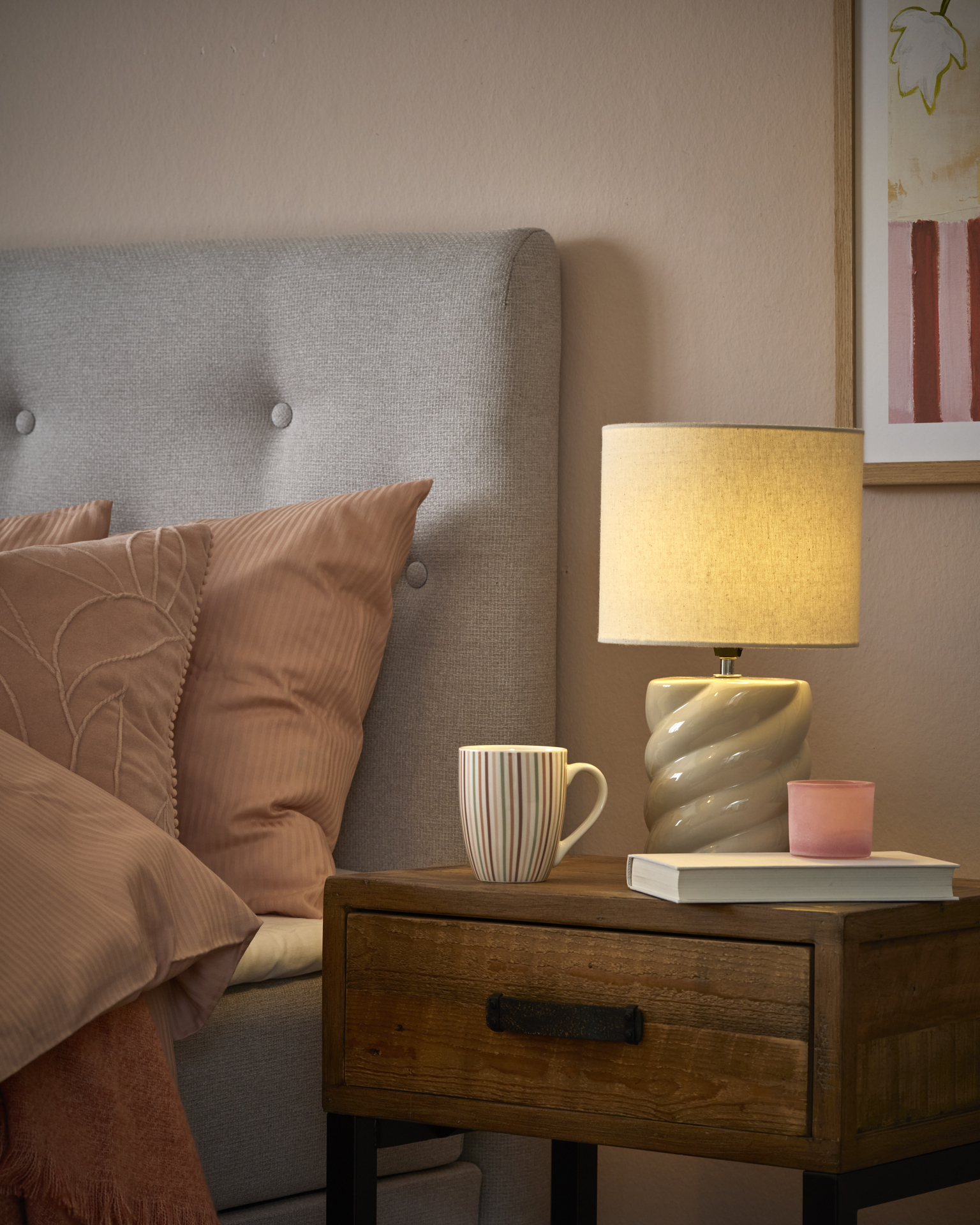 På ett sängbord i rustikt trä står en grå bordslampa och en randig kopp.