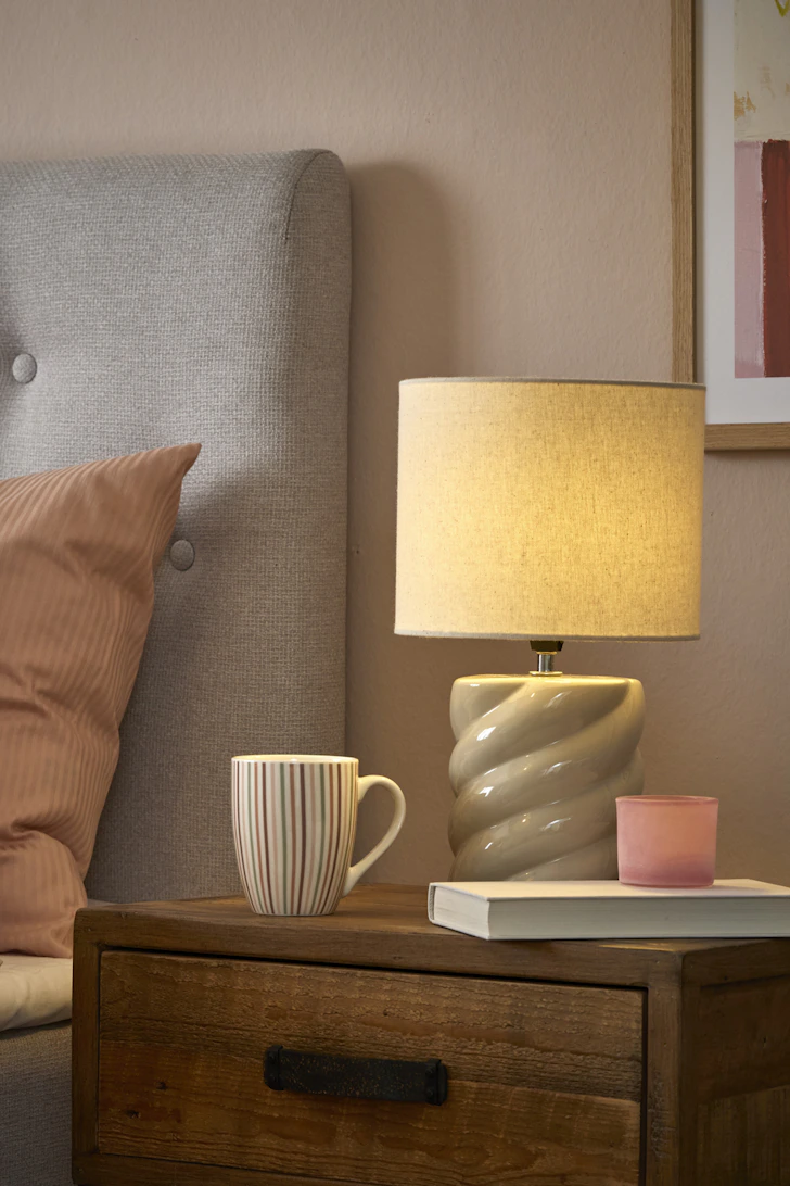På ett sängbord i rustikt trä står en grå bordslampa och en randig kopp.
