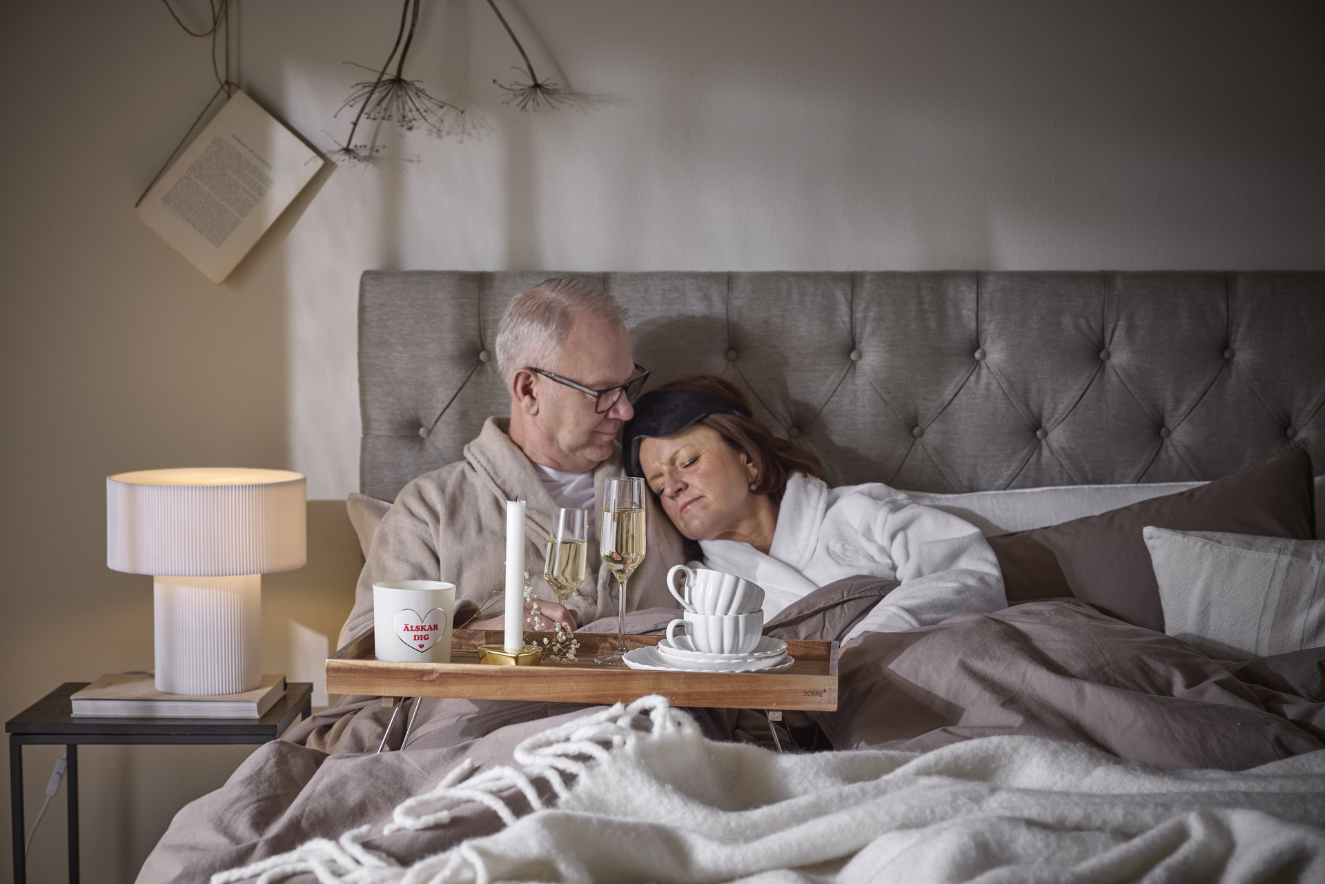 en man och en kvinna sitter i en säng med en bricka i knät där det står en ljus, champagneglas och muggar