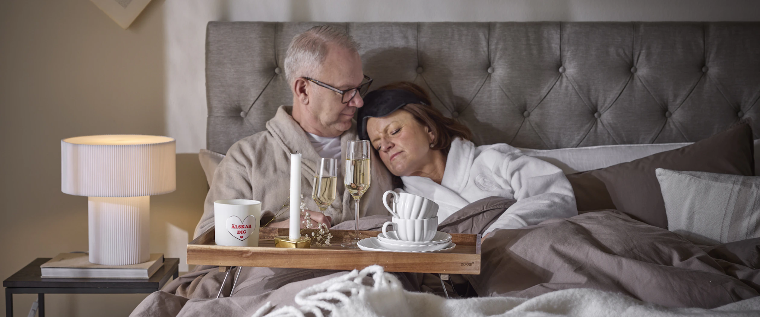 en man och en kvinna sitter i en säng med en bricka i knät där det står en ljus, champagneglas och muggar