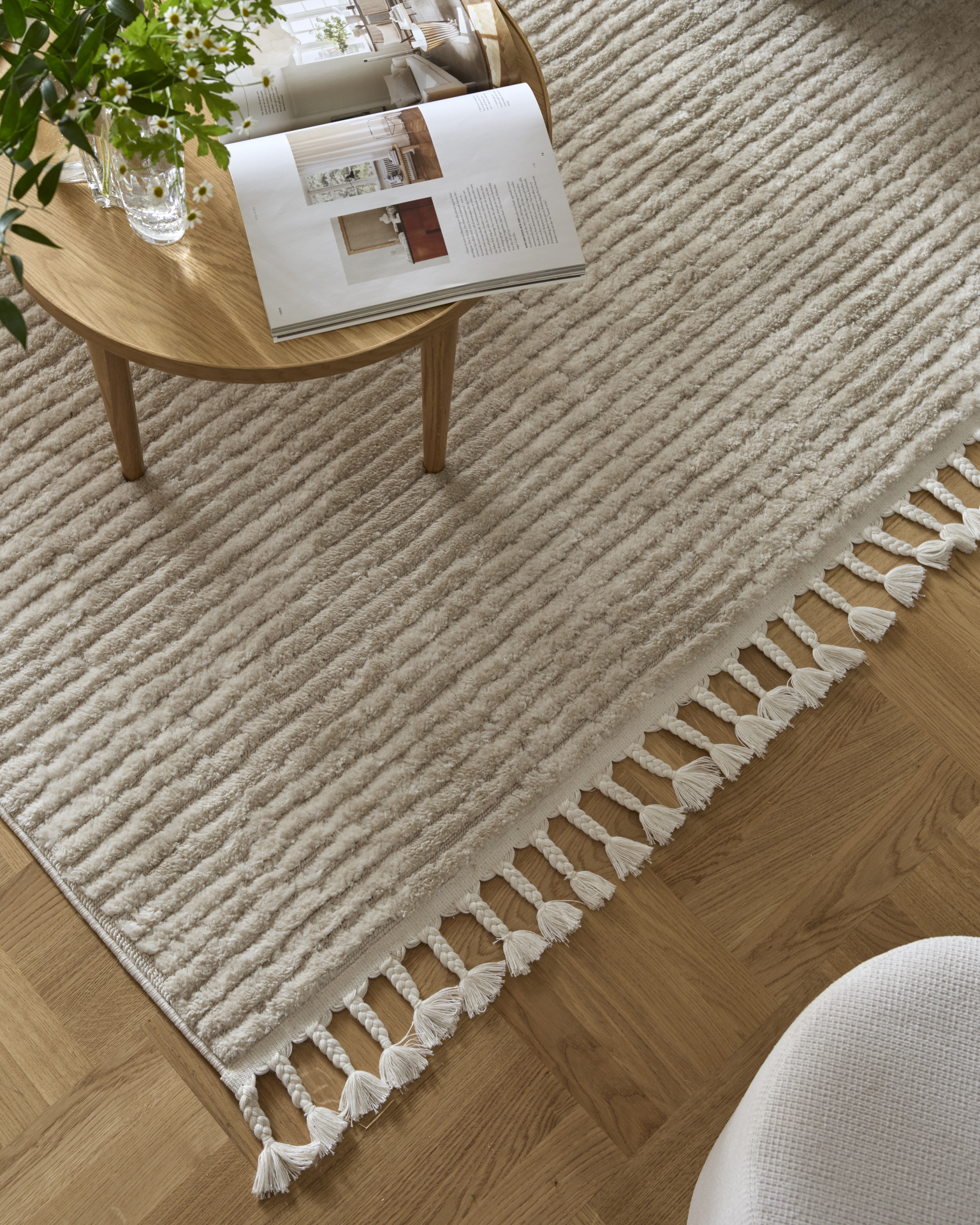 En vävd, vit matta med fransar ligger på ett ljust ekgolv, på mattan står ett soffbord i ek med en tidning uppslagen