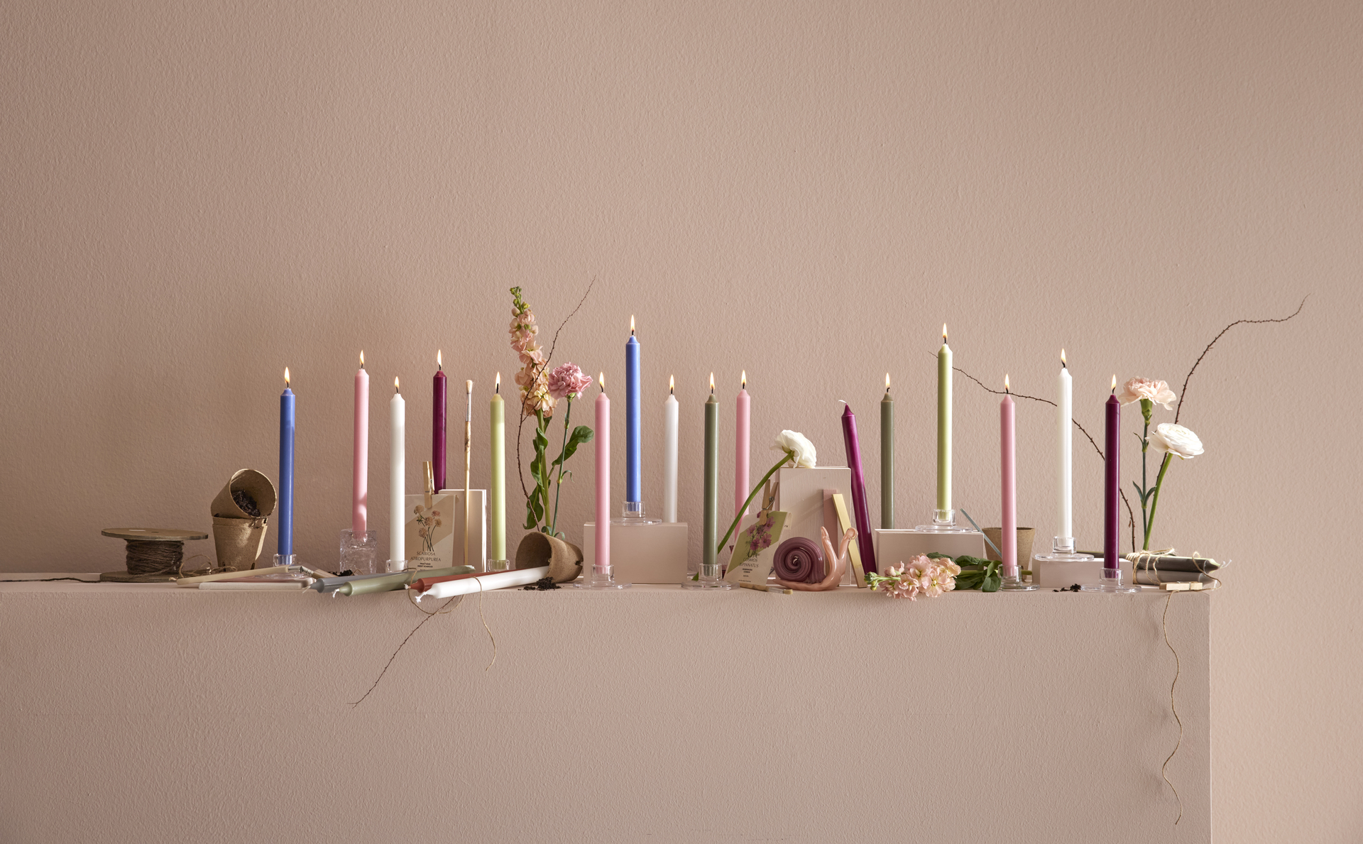 Flera rustikljus i olika färger står uppradade i ljusstakar tillsammans med vårblommor och planteringskrukor.