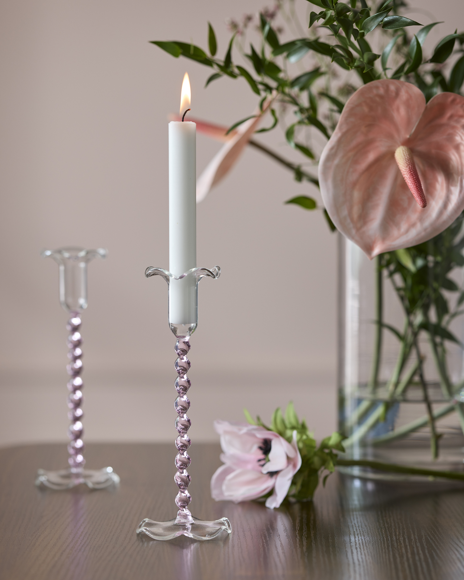 På ett bord i mörkt trä står två ljusstakar i rosafärgat glas intill en vas med rosa blommor