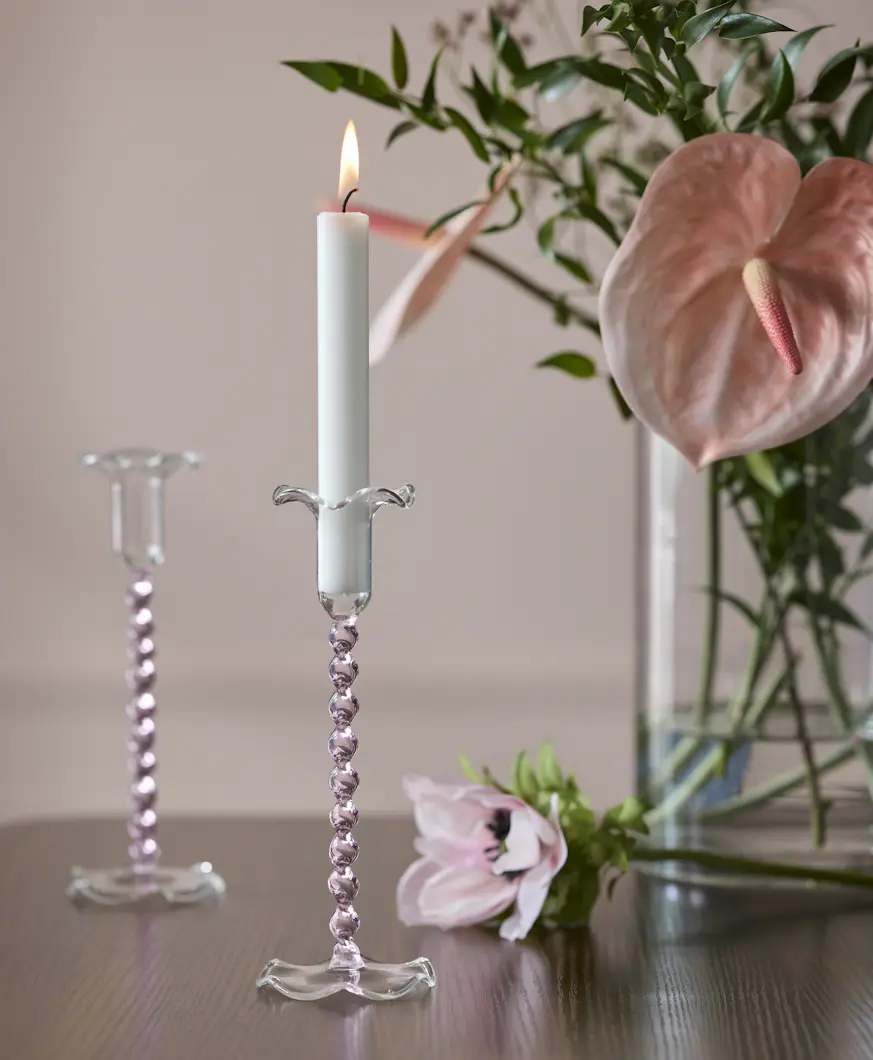 På ett bord i mörkt trä står en ljusstake i rosa glas framför en vas med rosa blommor.