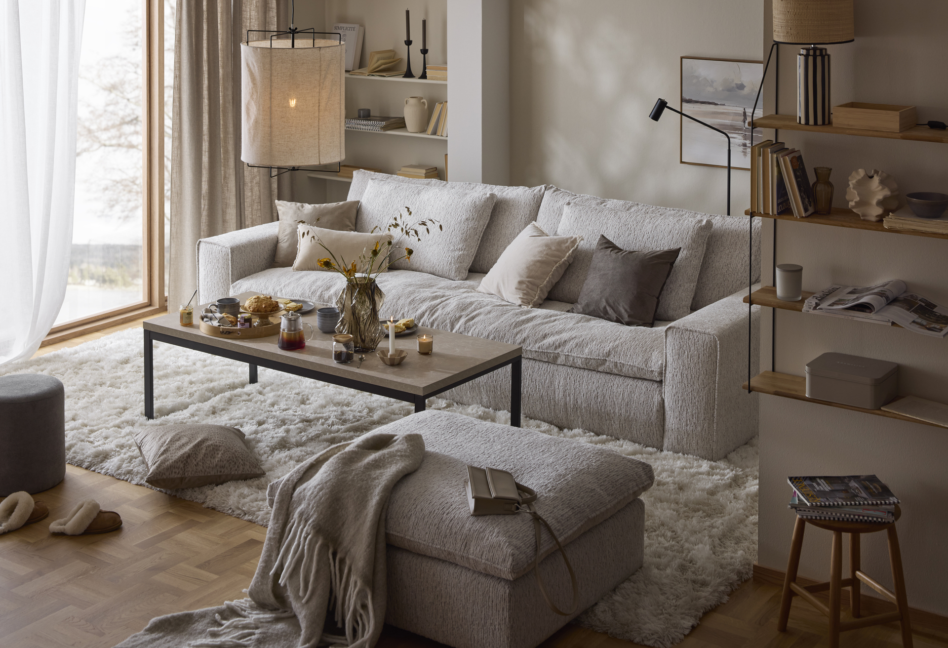 En grå stor soffa står i ett vardagsrum på en vit fluffig matta. Intill finns ett rektangulärt soffbord i sten och en beige fotpall. 