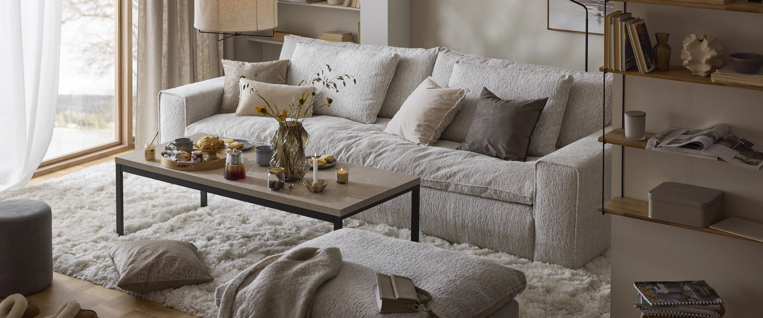 En grå stor soffa står i ett vardagsrum på en vit fluffig matta. Intill finns ett rektangulärt soffbord i sten och en beige fotpall.