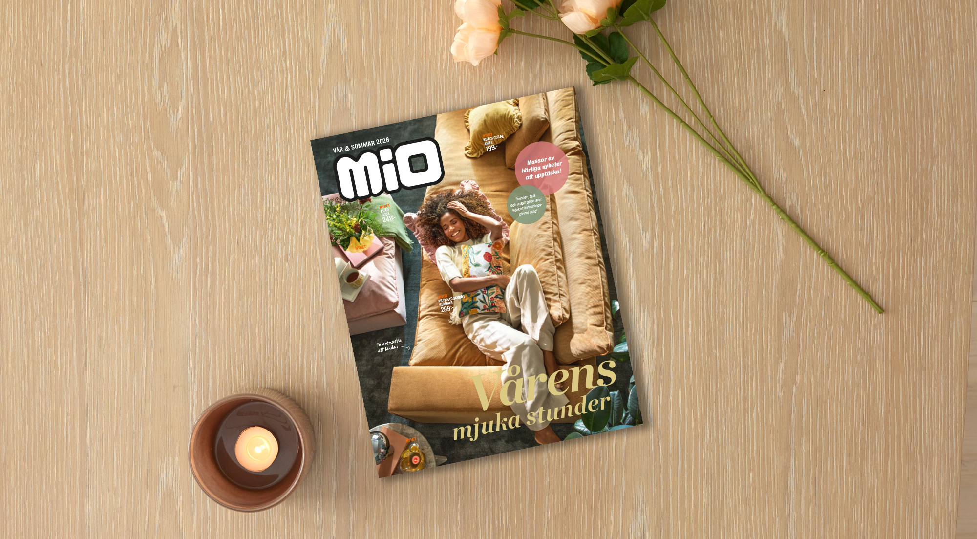 På ett ljust träbord ligger Mios vårkatalog 2026 intill ett ljus och en blomma.