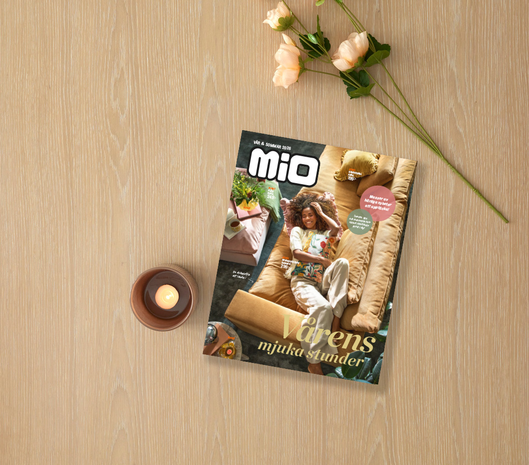 På ett ljust träbord ligger Mios vårkatalog 2026 intill ett ljus och en blomma.