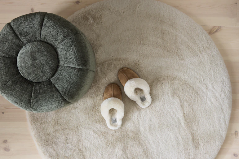 På en vit rund matta står en grön fotpall och ett par bruna tofflor.