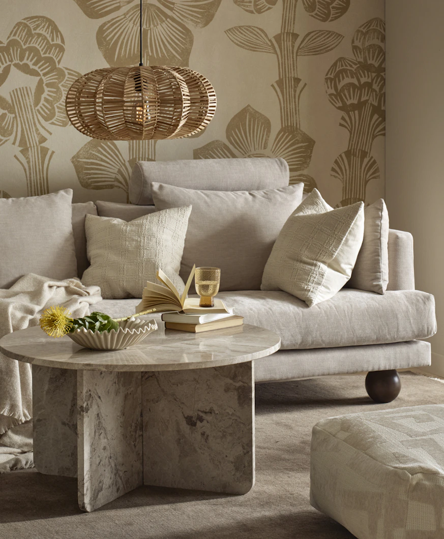 I ett vardagsrum med mönstrad, beige tapet står en beige soffa med klotformade ben