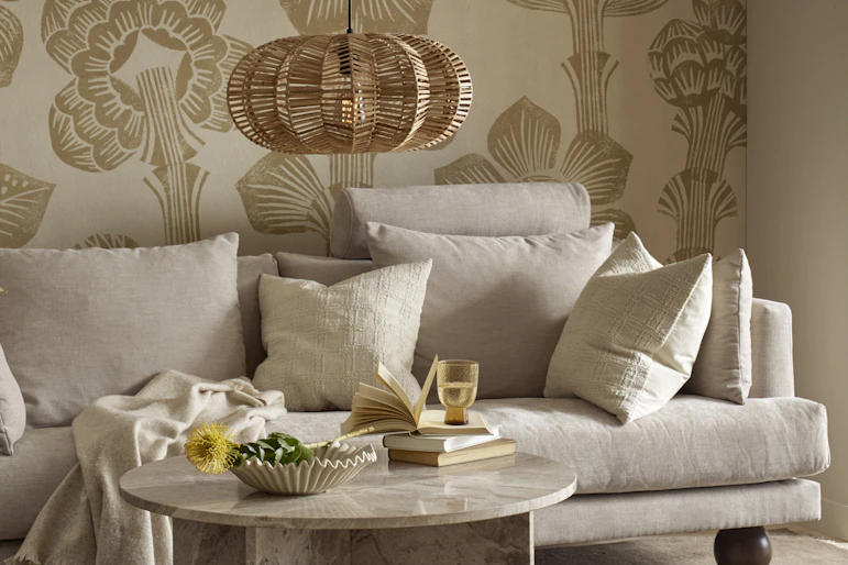 I ett vardagsrum med mönstrad, beige tapet står en beige soffa med klotformade ben