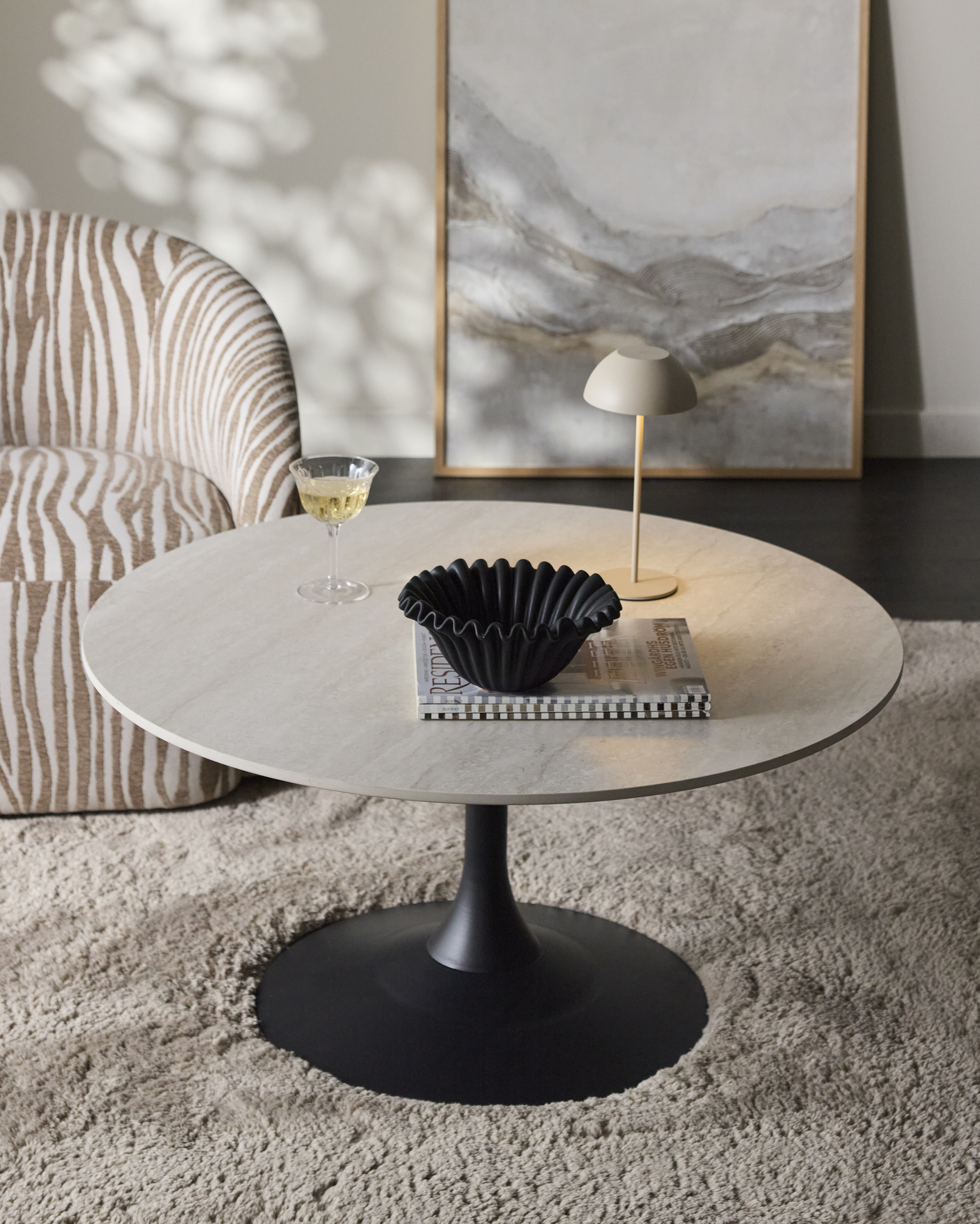 Ett runt soffbord med bordsskiva i sten och svart pelarfot står på en fluffig matta intill en fåtölj