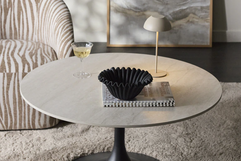 Ett runt soffbord med bordsskiva i sten och svart pelarfot står på en fluffig matta intill en fåtölj
