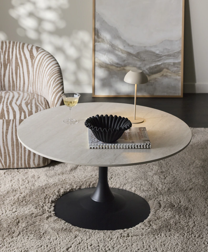 Ett runt soffbord med bordsskiva i sten och svart pelarfot står på en fluffig matta intill en fåtölj