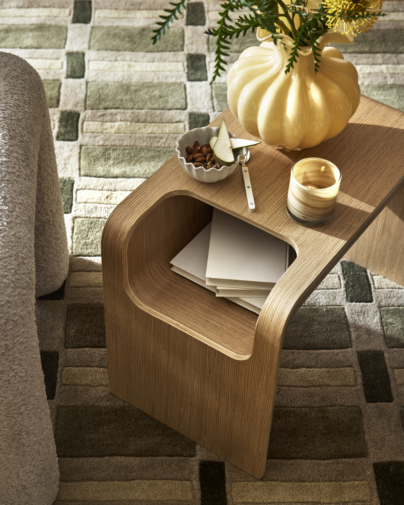 På en grön, mönstrad matta står ett sidobord i ek dekorerad med en gul vas och en ljuslykta