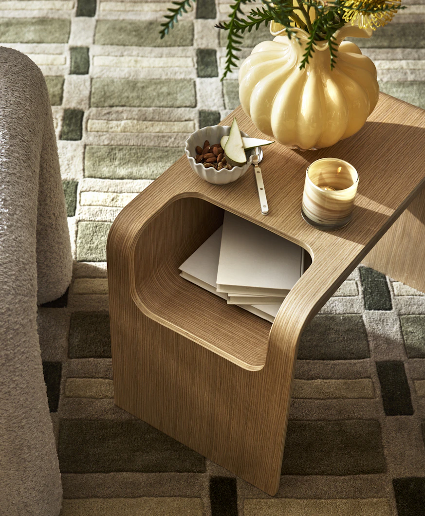 På en grön, mönstrad matta står ett sidobord i ek dekorerad med en gul vas och en ljuslykta