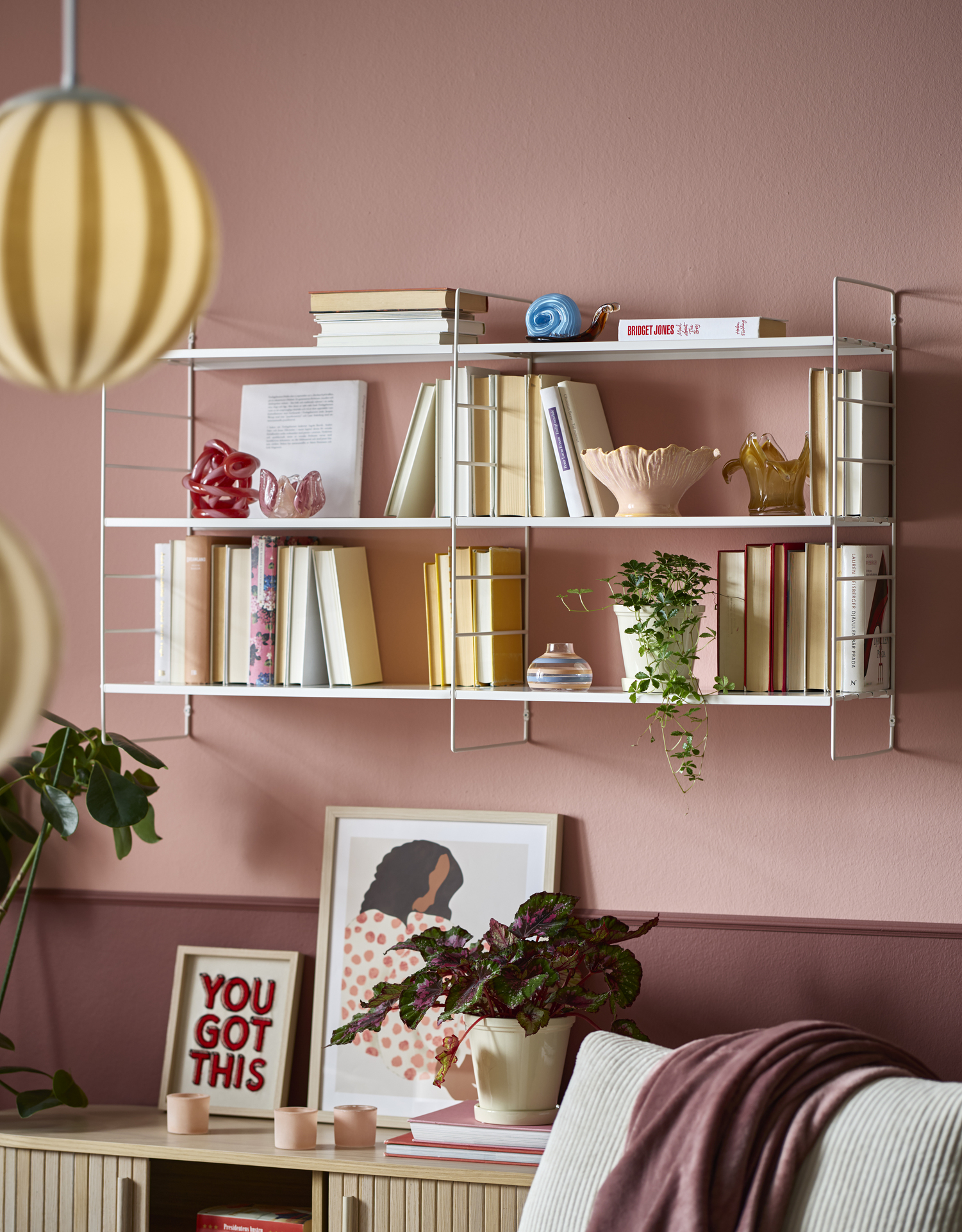 På en rosa vägg sitter en vit vägghylla full med böcker och dekoration