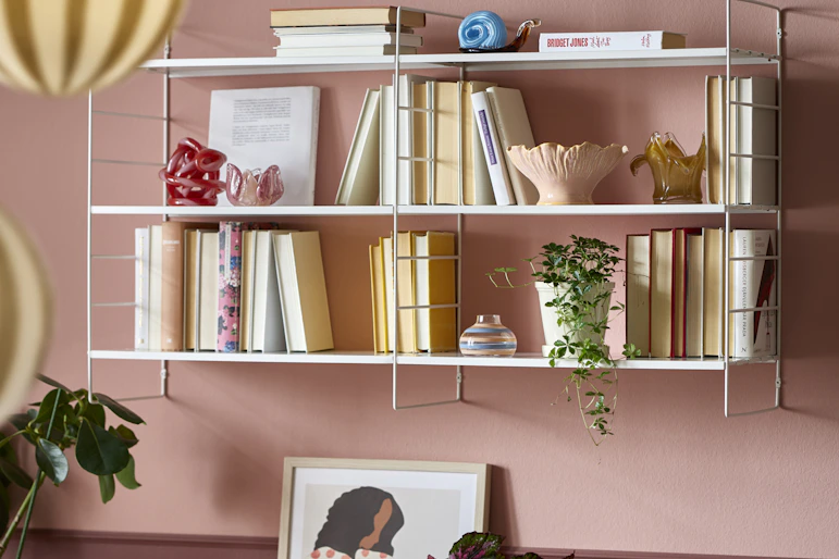 På en rosa vägg sitter en vit vägghylla full med böcker och dekoration