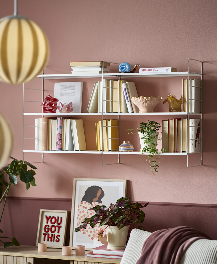 På en rosa vägg sitter en vit vägghylla full med böcker och dekoration