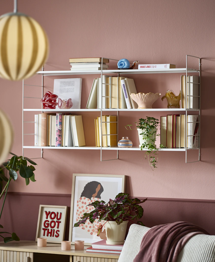 På en rosa vägg sitter en vit vägghylla full med böcker och dekoration