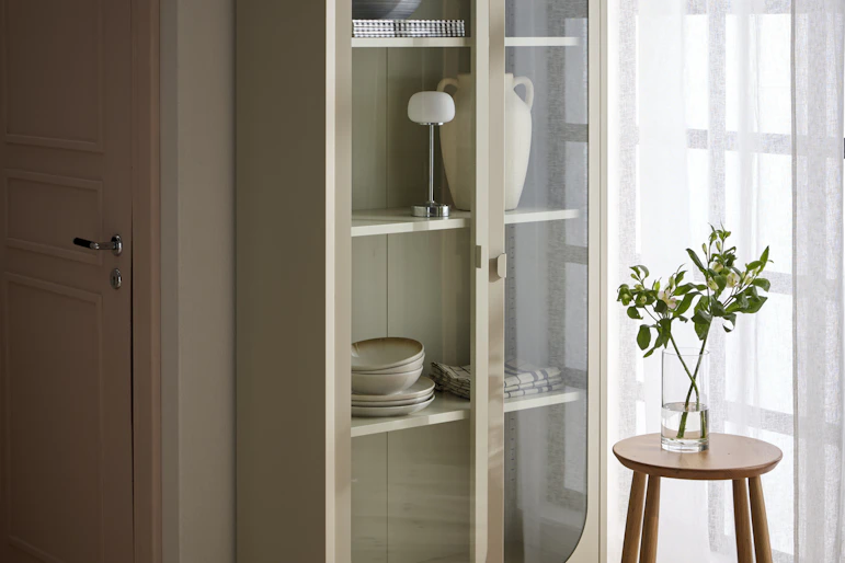 Ett beige vitrinskåp är fyllt med porslin, en urna och en lampa.